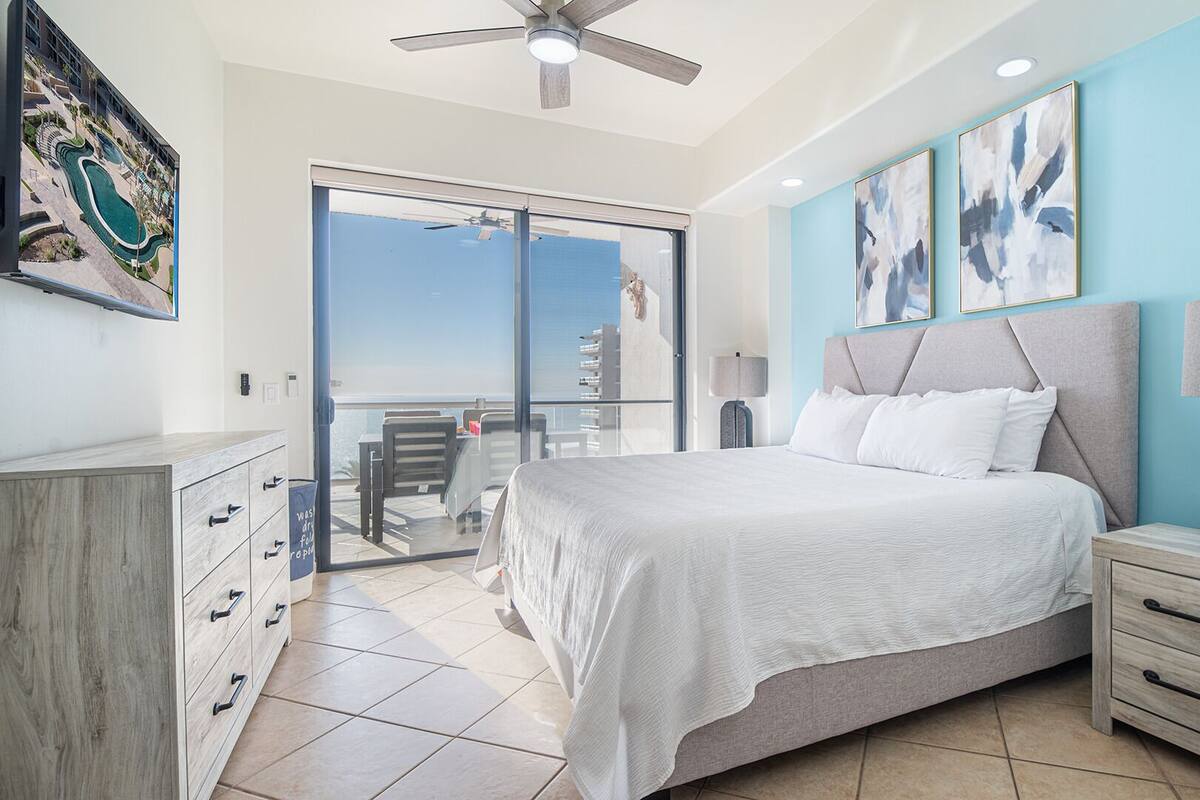 Une chambre à coucher comprenant un lit, une table de chevet, une lampe, une commode et une vue sur une piscine et un immeuble, visible à travers une porte-fenêtre coulissante.
