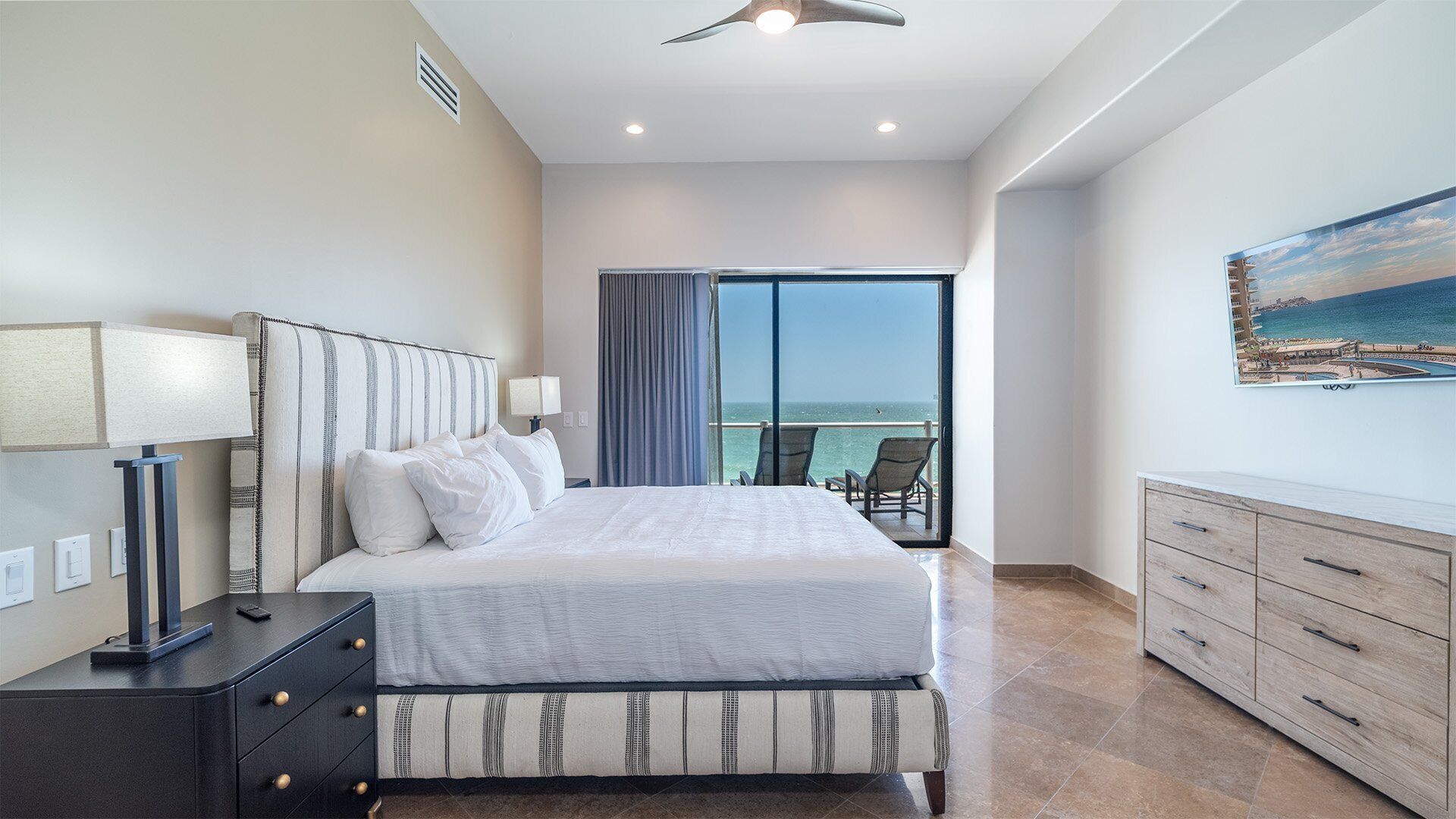 A modern hotel room with a large bed, a desk, a chair, and a balcony with a view of the sea.