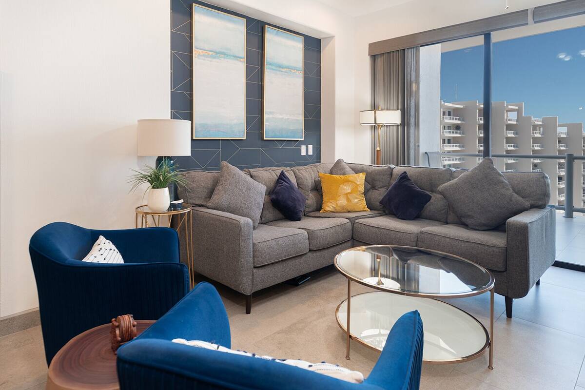 Ein modernes Wohnzimmer mit einem grauen Ecksofa, einem blauen Sessel und einem Glastisch. Ein Bücherregal mit Dekorationselementen und ein großes Fenster mit Blick auf Gebäude.