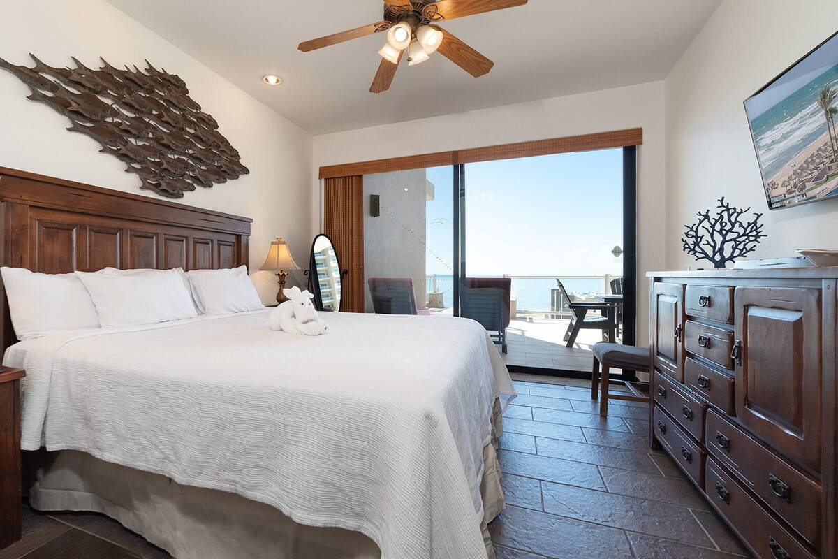 Une chambre d’hôtel avec un lit, des tables de chevet en bois, une commode, un ventilateur de plafond, une grande fenêtre et une vue sur l’océan.