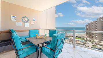 Un balcón con sillas turquesas y una mesa, con vista a una playa y edificios de un hotel.