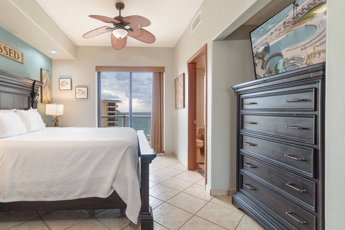 Une chambre d’hôtel avec un lit, un ventilateur de plafond, une vue sur l’océan et une grande commode en bois sombre.