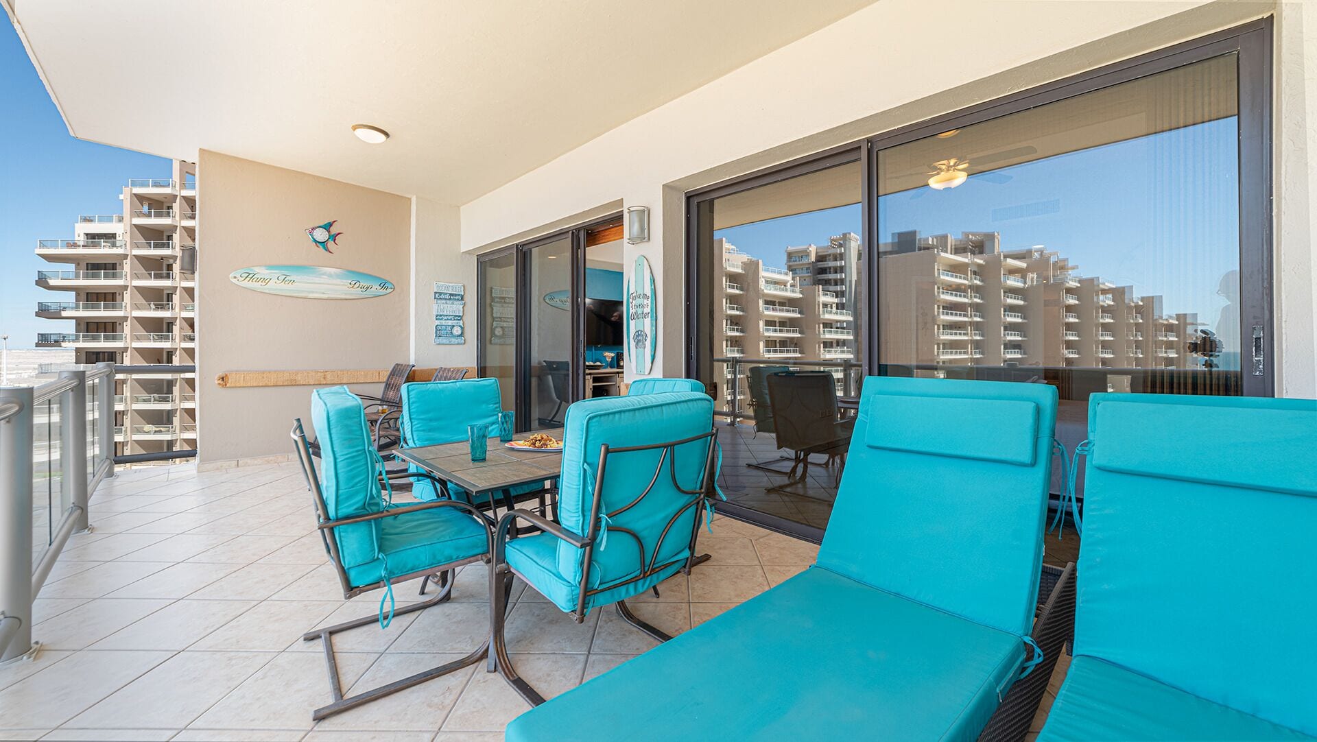 A balcony with turquoise chairs and a table, large windows with a city view, and a surfboard decoration on the wall.