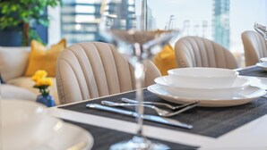 Un área de comedor con una mesa preparada para dos personas, una copa de martini y vistas a la ciudad a través de amplias ventanas.