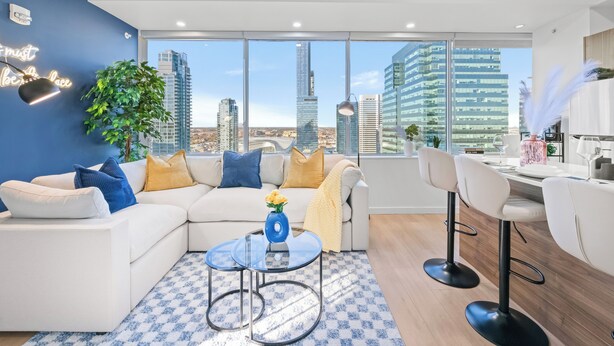 Una sala moderna con un sofá blanco, cojines azules y amarillos, una mesa de centro de vidrio con un jarrón de flores amarillas, y un ventanal con vistas a la ciudad.