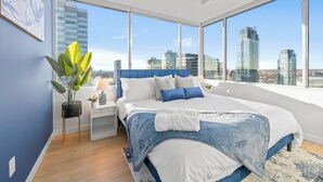 Un dormitorio moderno con una cama grande, una mesita de noche, una planta en maceta y vistas a la ciudad a través de amplios ventanales.