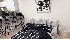 Un dormitorio moderno con cama, una zona de cocina con armarios blancos, una mesa de comedor con sillas y un aire acondicionado instalado en la pared.