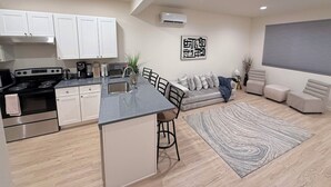 Cocina moderna con una isla central, armarios blancos y electrodomésticos de acero inoxidable. Cuenta con una zona de estar con sofá y sillones, un tapete estampado y una obra de arte colgada en la pared.