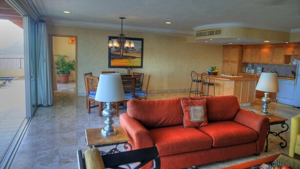 A living room with a red sofa, a dining area with chairs, a kitchen with wooden cabinets, and a large window with curtains. - 3-bedroom Ocean Front PENTHOUSE in marvelous Cabo San Lucas (Cabo San Lucas)