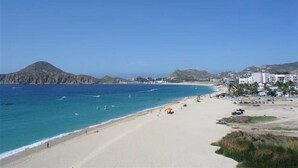 A beach with clear blue water, a sandy shore, and buildings in the background. - 3-bedroom Ocean Front PENTHOUSE in marvelous Cabo San Lucas (Cabo San Lucas)