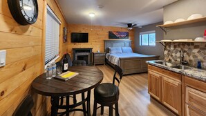 A cozy cabin interior with a wooden wall clock, a fireplace, a bed, a dining table with chairs, and a kitchen area. - Elk Haven Cabins - Cabin 4 (Payson)