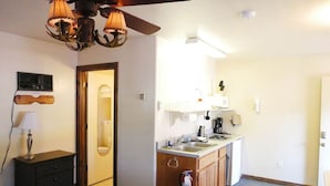Ett kompakt hotellrum med ett litet pentry, en takfläkt, ett nattduksbord med en lampa och en dörr som leder till badrummet.