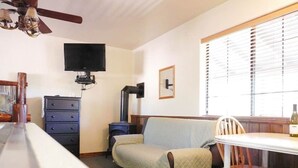 A compact room with a sofa, a small table, a TV mounted on the wall, and a window with blinds. - Elk Haven Cabins - Cabin 2 (Payson)