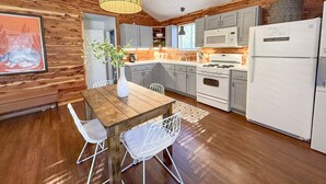 A modern kitchen with wooden walls, a dining area with a wooden table and white chairs, and a hanging light fixture. - Skip Rock Cottage ~ ON Livingston Creek (Mountain View)