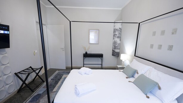 Ett hotellrum med en säng med himmelssäng, ett litet skrivbord med en lampa, en tv som är monterad på väggen och en mönstrad gardin.