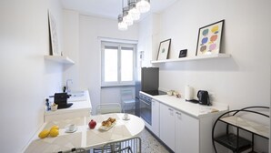 Ett kompakt kök med vita skåp, ett matbord med stolar, en inbyggd ugn och en hängande lampa.