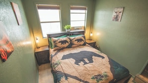 A bedroom with a bed, two windows, a nightstand, a lamp, and a wall decoration. - Paula's Place Near The Lake (Elephant Butte)