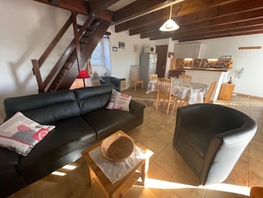 Ein Wohnzimmer mit schwarzem Sofa, einem Holztisch, einem Essbereich mit Tisch und Stühlen sowie einer Küche im Hintergrund.