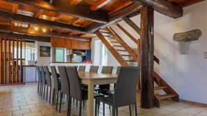 Un espace repas avec une longue table et des chaises, un escalier en bois et une cuisine en arrière-plan.