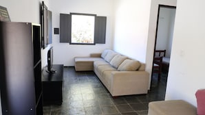 Un salon avec un canapé d’angle beige, un meuble de télévision noir et un écran de télévision, ainsi qu’une fenêtre avec des stores.
