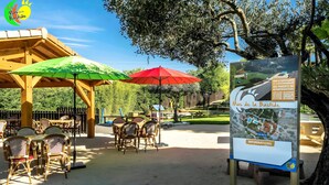 Un espace extérieur aménagé avec des tables, des chaises, des parasols et un panneau d’affichage.