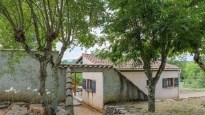 Un bâtiment rustique avec un toit en tuiles, entouré d’arbres et d’un mur de pierres.