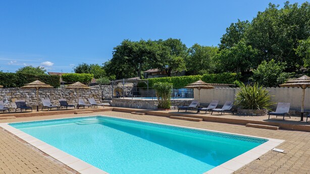 Une piscine rectangulaire bordée de carreaux, entourée de chaises longues et de parasols, avec un mur de pierre et de la verdure en arrière-plan.