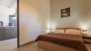 Une chambre à coucher comprenant un lit, des tables de chevet, une commode, un tableau accroché au mur et donnant sur la cuisine adjacente.