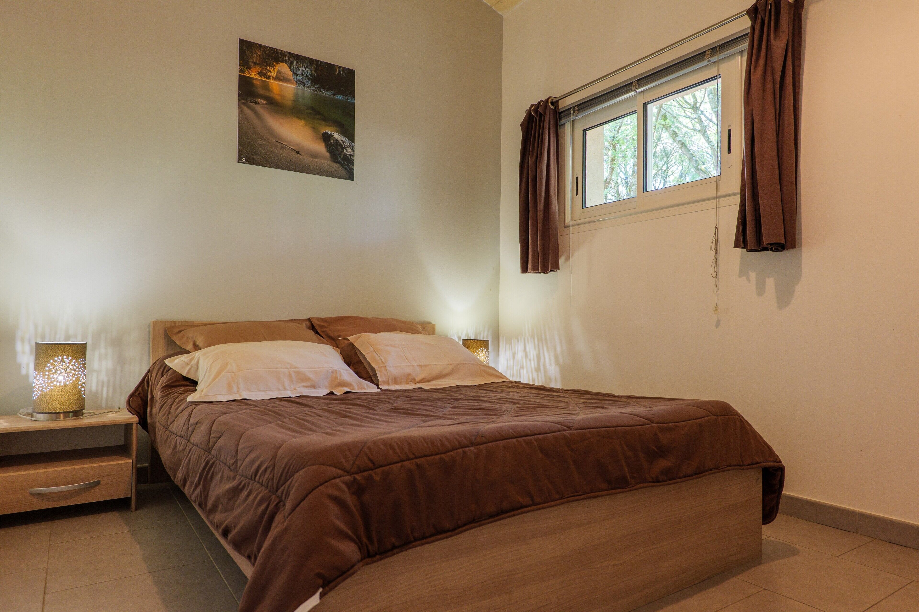Une chambre à coucher avec un lit, une table de chevet, une lampe, une fenêtre avec des rideaux et un tableau au mur.