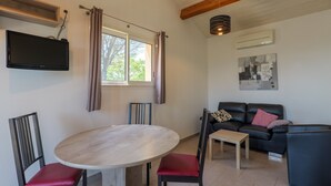 Un salon avec une table ronde, deux chaises, un canapé, un téléviseur fixé au mur et une fenêtre avec des rideaux.