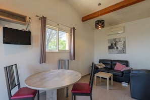 A living room with a round table, two chairs, a sofa, a TV mounted on the wall, and a window with curtains.