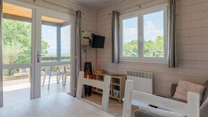 A room with a wooden floor, a white table, a sofa, a TV mounted on the wall, and a window with curtains. - Gîte Romarin 1 – Village Holiday Rentals (Joyeuse)