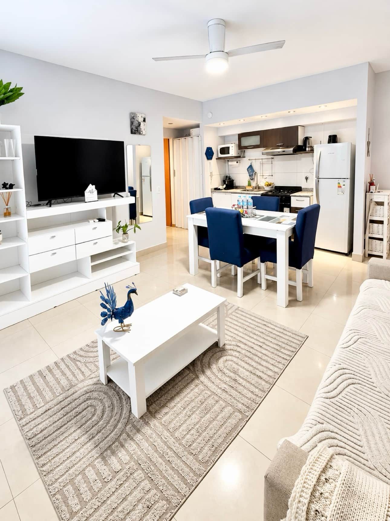 A modern living room with a flat-screen TV, a dining area with blue chairs, a kitchen with white appliances, and a ceiling fan.