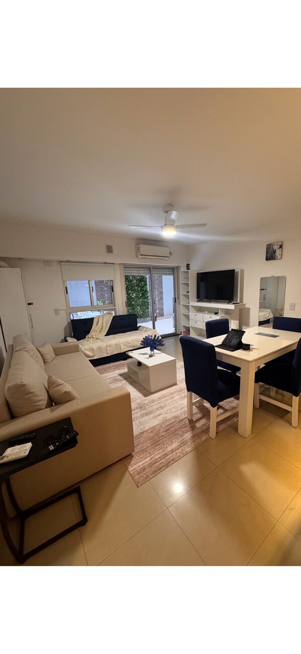 A modern living room with a sofa, a dining table with chairs, a flat-screen TV, and a large window.