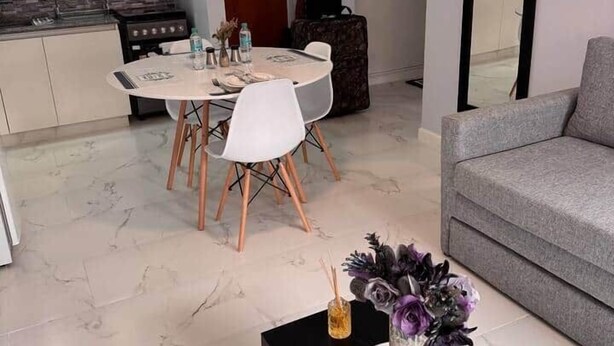 Una sala moderna con un sofá gris, un comedor con una mesa redonda y sillas, una cocina con gabinetes blancos y salpicadero de azulejos, y una mesa de centro negra con un jarrón.