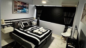 Un dormitorio con cama, escritorio, silla, ventilador de techo, ventana con cortinas y un cuadro en la pared.
