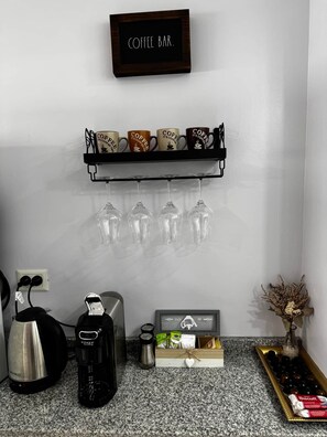 A coffee bar area with a shelf holding coffee mugs and glasses, a coffee maker, a kettle, and a small plant.