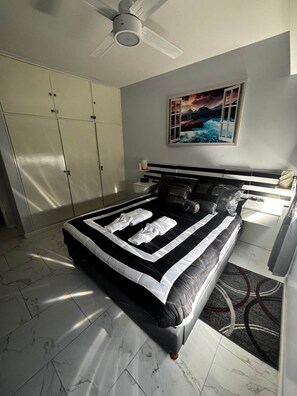A bedroom with a bed, a ceiling fan, a wardrobe, a painting, and a nightstand.