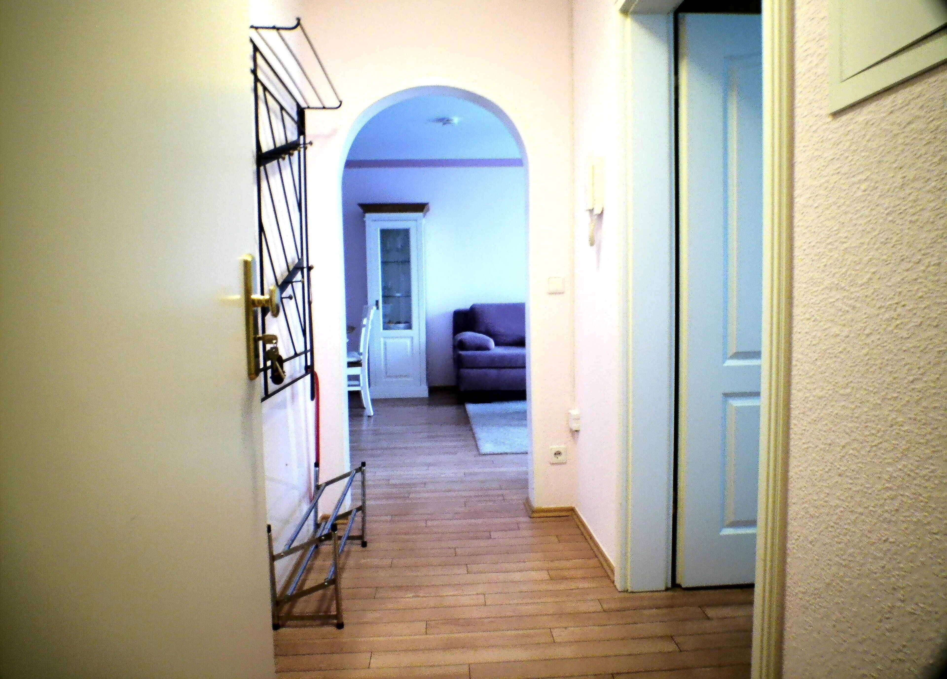 Um corredor com piso de madeira, uma porta branca à direita e um sofá roxo ao fundo.