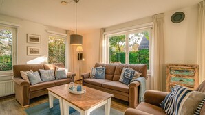 A cozy living room with two sofas, a coffee table, and a lamp. There are windows with curtains and a view of greenery outside. - Charming Villa Near the sea and Forest (De Koog)