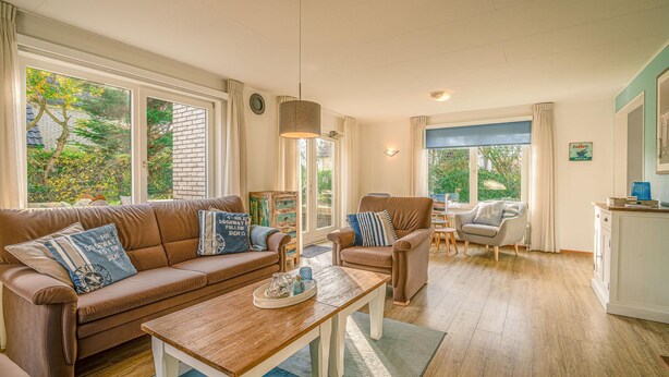 A cozy living room with a brown sofa, a wooden coffee table, and a chair. There are large windows with curtains, a lamp, and a cabinet. - Charming Villa Near the sea and Forest (De Koog)