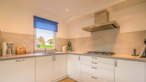 A modern kitchen with white cabinets, a gas stove, and a window with a blue blind. - Charming Villa Near the sea and Forest (De Koog)
