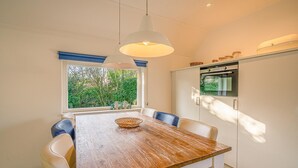 A dining area with a wooden table, white chairs, a hanging light fixture, and a view of greenery through a window. - Charming Villa Near the sea and Forest (De Koog)
