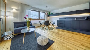A modern living room with a sofa, a round table, and a coffee table. There is a kitchen area with dark cabinets and a window overlooking a cityscape. - Apartment with a view of the surrounding mountains (Pfalzen)