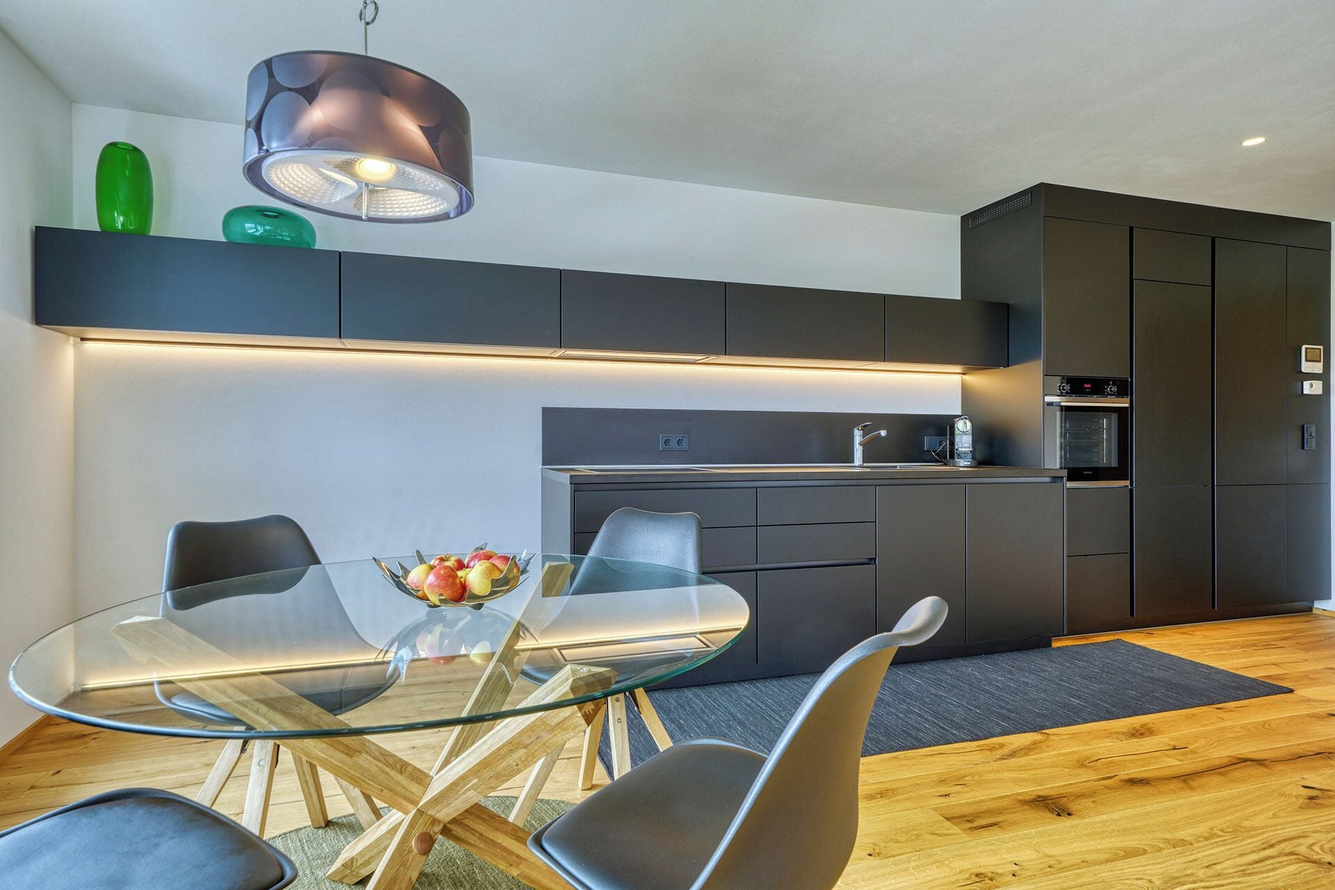 Une cuisine moderne avec des meubles de couleur sombre, une table à manger avec un plateau en verre et un four encastré.