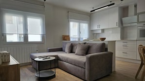 Ein modernes Wohnzimmer mit grauem Sofa, einem kleinen runden Tisch und einer Küchenzeile mit weißen Schränken.