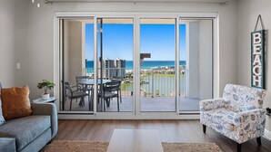 A living room with a sofa, armchair, and a dining table with chairs. A large sliding glass door leads to an outdoor balcony with a view of the beach and ocean. - Ariel Dunes II 1305 (Miramar Beach)