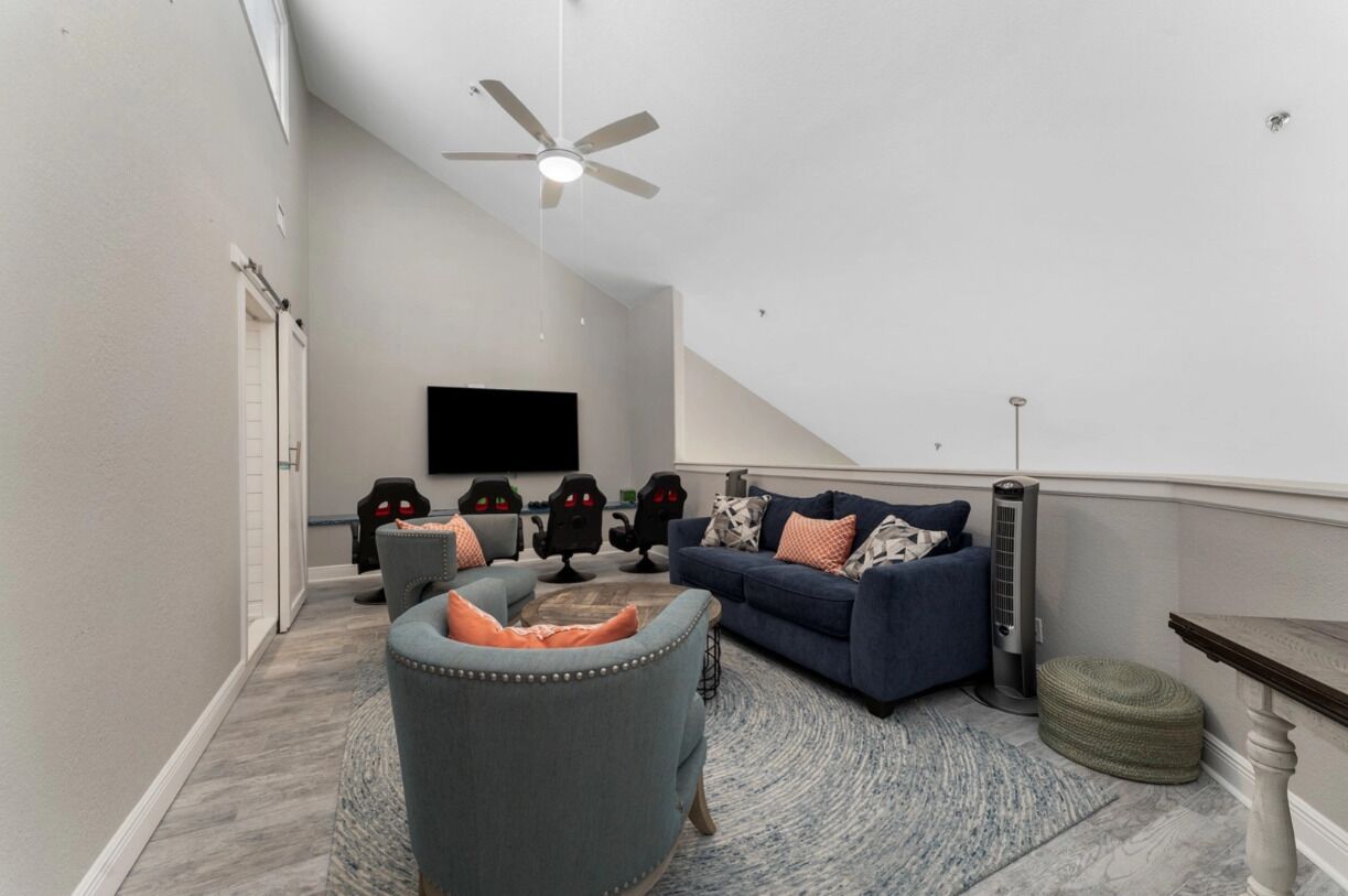 A modern living room with a ceiling fan, a flat-screen TV mounted on the wall, a blue sofa with orange cushions, a grey armchair, and a small round ottoman.