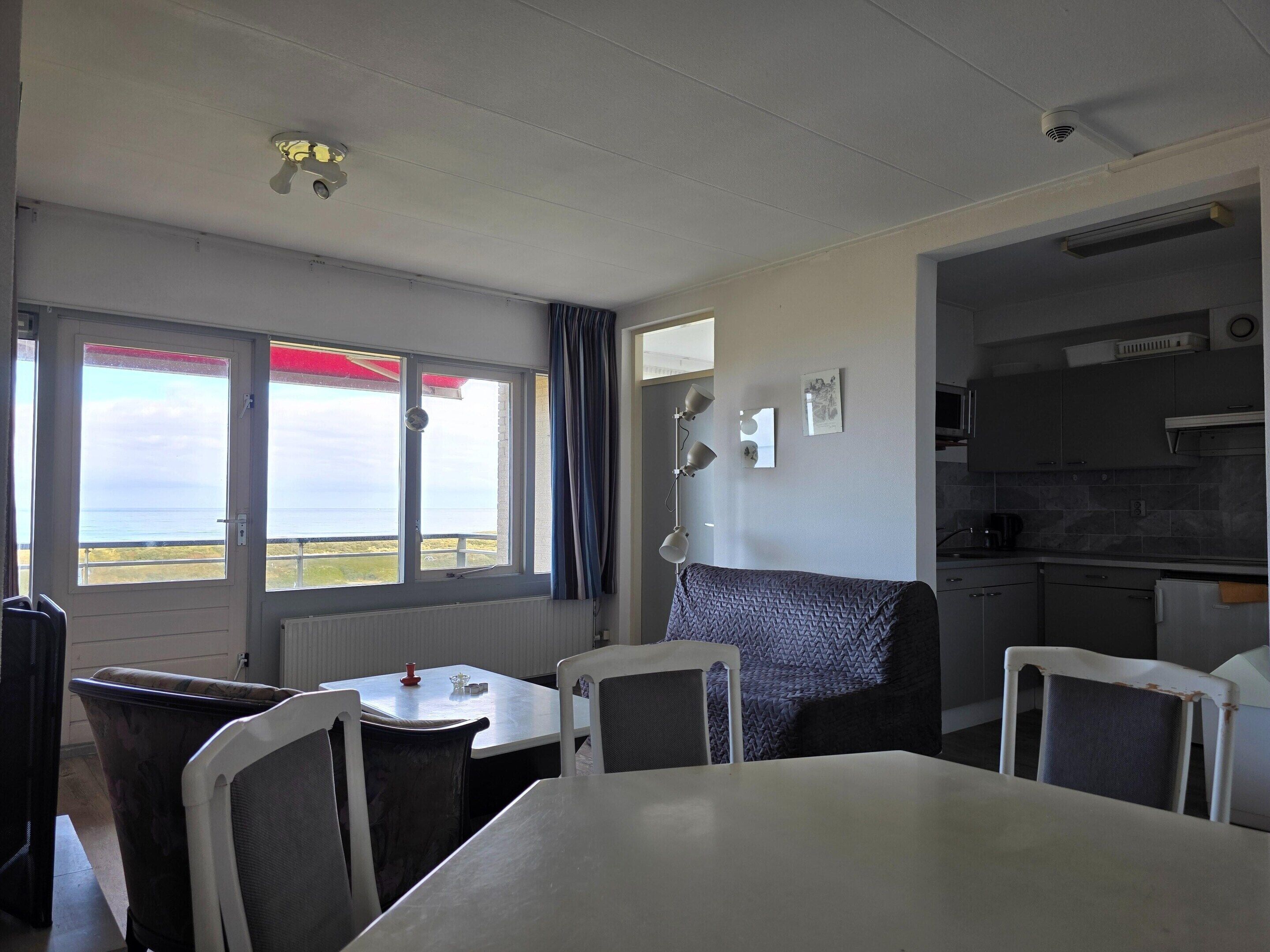 A room with a dining table, chairs, a view of the sea through large windows, a kitchenette, and a ceiling fan.