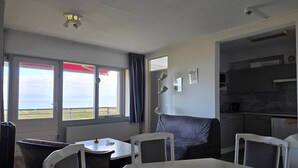 A room with a dining table, chairs, a view of the sea through large windows, a kitchenette, and a ceiling fan. - 2 Person Apartment With sea View (De Koog)